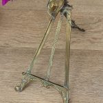 Antique Western style brass tabletop easel featuring a detailed miniature cowboy hat cresting on a folding tripod frame with natural green verdigris patina.