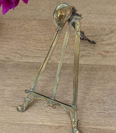 Antique Western style brass tabletop easel featuring a detailed miniature cowboy hat cresting on a folding tripod frame with natural green verdigris patina.